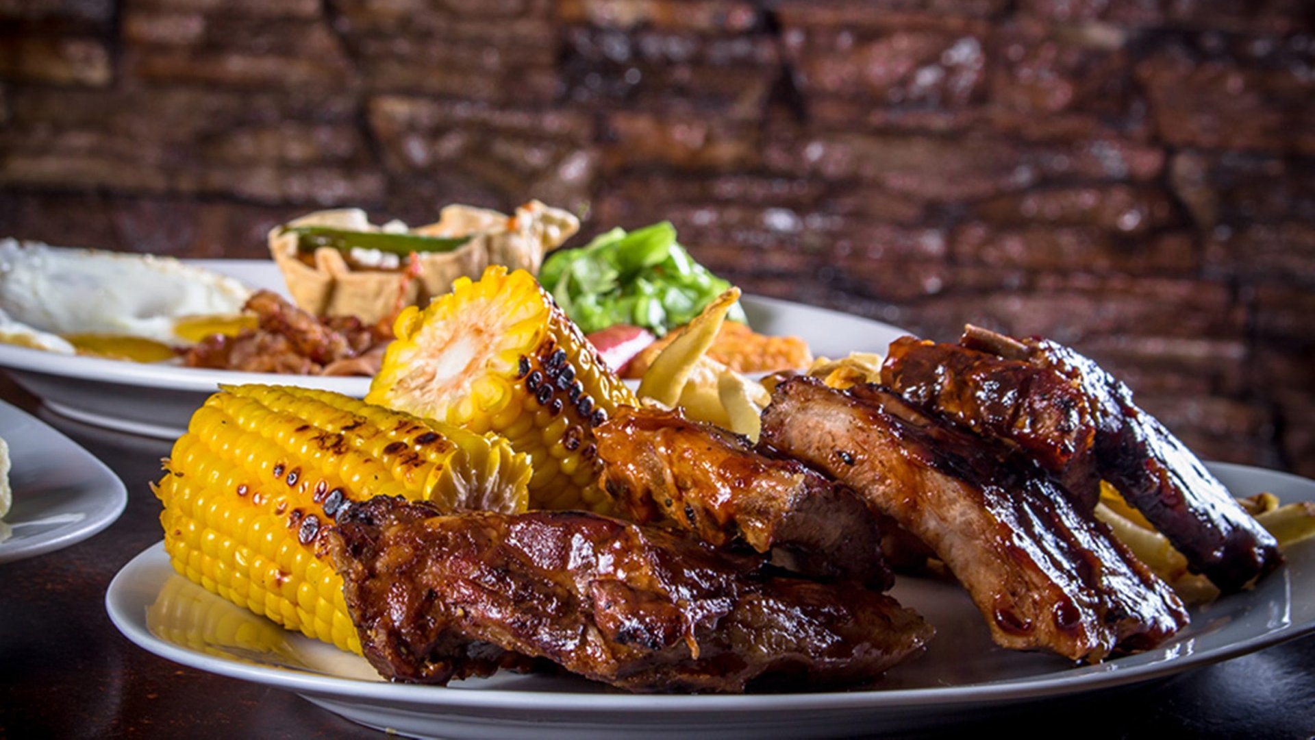 Costillas a la parrilla con elote asado, servidas frente al muro de ladrillo del salón